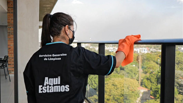 Personal uniformado de Aquí Estamos limpiando la baranda del balcón de una torre corporativa, con la ciudad de fondo